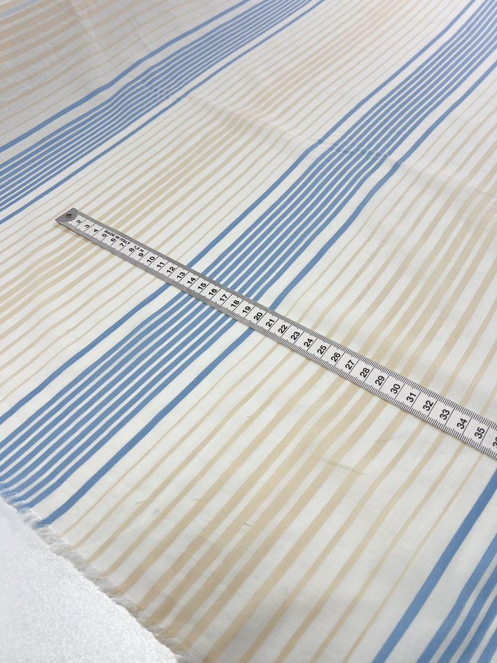 A close-up image of GOTS Cotton - Buttercream Stripe from Super Cheap Fabrics, showcasing alternating vertical stripes in blue and beige. A measuring tape is laid diagonally across the 160cm organic cotton fabric, displaying measurements in centimeters and emphasizing its eco-friendly fashion appeal.