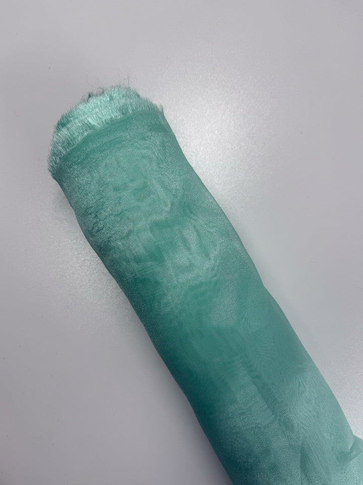 A rolled-up piece of Super Cheap Fabrics’ Organza - Aqua Glass - 150cm, in light teal with a soft, shiny texture, lies on a smooth white surface. A hint of sheer organza adds delicate elegance; the satin edge is slightly frayed.