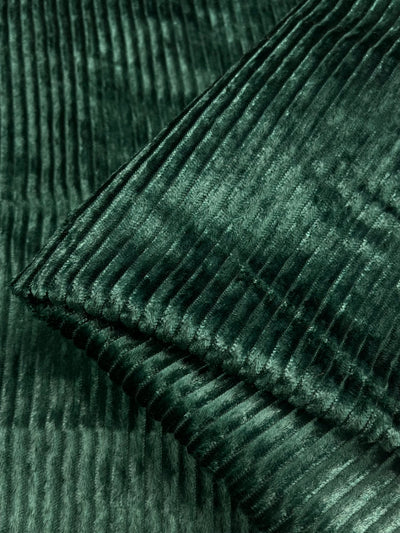 A close-up of Super Cheap Fabrics' Velvet Medium Cord in Forest Biome showcases its textured, ribbed pattern. The dark green fabric appears soft and luxurious with a slight sheen reminiscent of heavyweight velvet. The edges are folded over, highlighting the thickness and texture of this 150cm wide material.