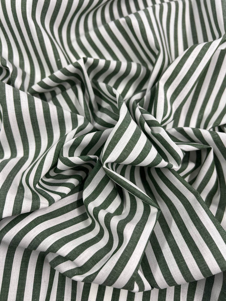 A close-up of the "Cotton Lawn - Fir - 150cm" from Super Cheap Fabrics shows gathered folds of green and white striped lightweight cotton. The alternating vertical stripes create a dynamic, textured pattern. The overall effect is both clean and detailed, showcasing the 100% cotton fabric's design.