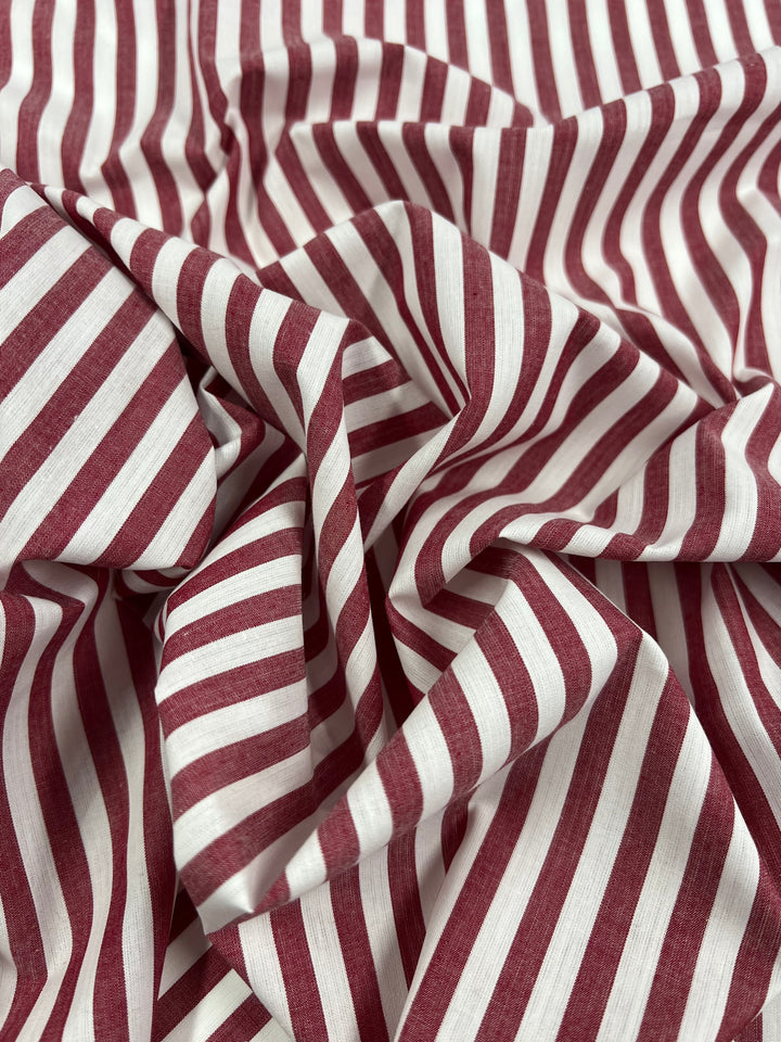 A close-up image of Super Cheap Fabrics' Cotton Lawn - American Beauty - 150cm. The red and white vertically striped fabric is lightweight, slightly wrinkled, and draped, showcasing variations in the stripe pattern and texture. Ideal for warm weather clothing, this material has a subtly silky finish.