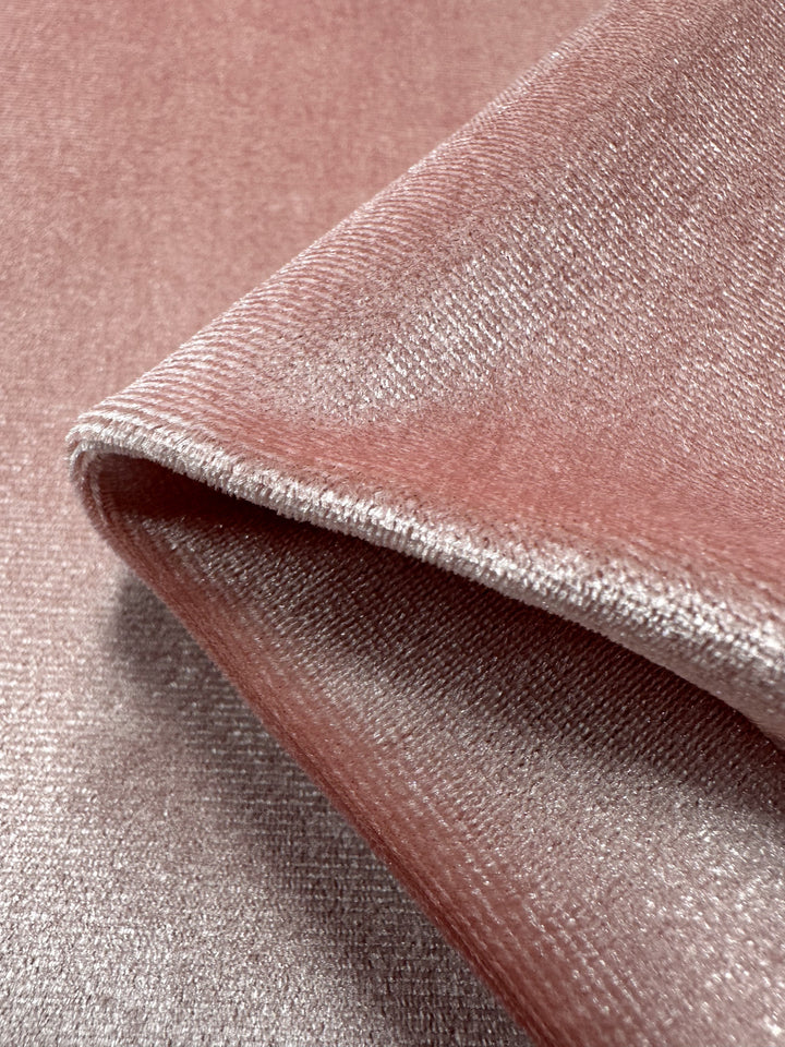 A close-up of the Stretch Velour - Peachskin from Super Cheap Fabrics shows a folded piece with a pink shimmering hue. Its texture looks soft and smooth, subtly reflecting light with a gentle sheen.