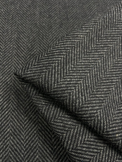 Close-up of Super Cheap Fabrics' Designer Wool - Herringbone - 150cm. The image shows a neatly folded section of this lightweight grey herringbone patterned fabric on another unfolded portion, highlighting the intricate zigzag weave design in a fine, textured material crafted in Italy.