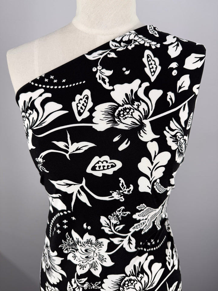 Super Cheap Fabrics’ Printed Lycra - Monochrome Botanica (150cm) is a black polyester spandex with bold white floral and leaf designs, shown draped asymmetrically over one shoulder on a dress form.