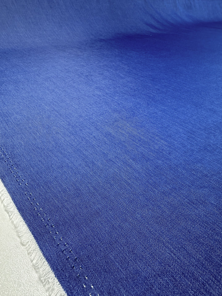 A close-up of a roll of Chambray - Victoria Blue fabric from Super Cheap Fabrics, featuring frayed edges on the bottom left. The detailed texture and weave are visible, highlighting its smooth, slightly glossy finish. The fabric extends into the background, indicating a large continuous piece.