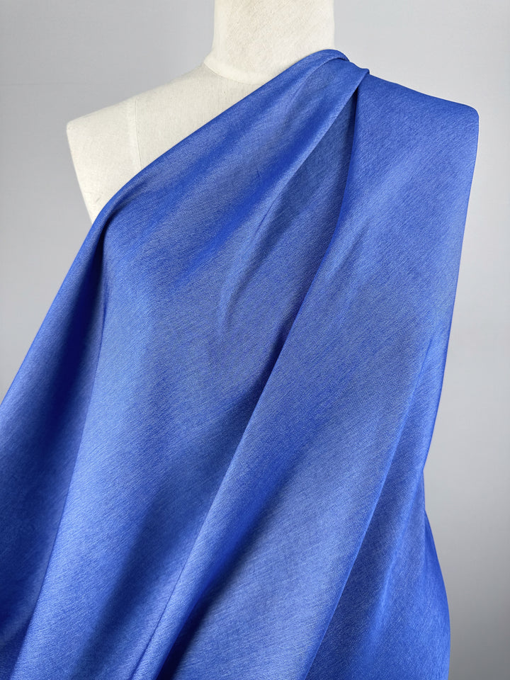 The Chambray - Victoria Blue - 150cm fabric from Super Cheap Fabrics elegantly drapes over the mannequin, its smooth texture gracefully wrapped over one shoulder. The striking Victoria Blue hue beautifully contrasts with the neutral background, highlighting its lightweight and graceful appearance.