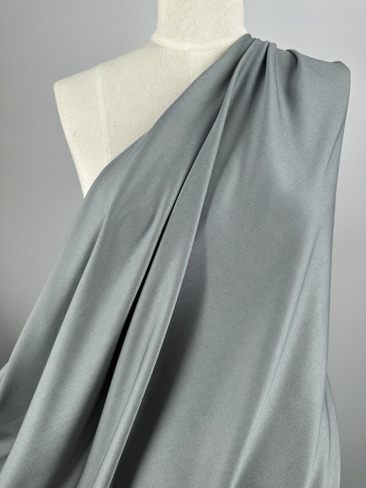 A mannequin draped with Super Cheap Fabrics' Chambray in Flint Grey - 150cm. The lightweight chambray fabric elegantly folds across one shoulder, creating gentle, flowing lines for a sophisticated look. The neutral light gray background complements the tone of the flint grey fabric beautifully.