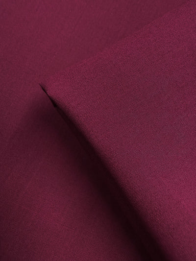 A close-up of Super Cheap Fabrics’ Poplin - Mulberry Matte (114cm) shows a rich maroon hue with visible weave texture; a folded corner highlights the smooth, matte finish and lightweight quality of this fabric.