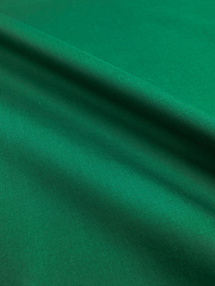 A close-up of Super Cheap Fabrics' Poplin - Emerald Plain (114cm) shows its smooth, vibrant emerald green surface with a diagonal fold casting a soft shadow, highlighting the fabric’s even texture and rich color.