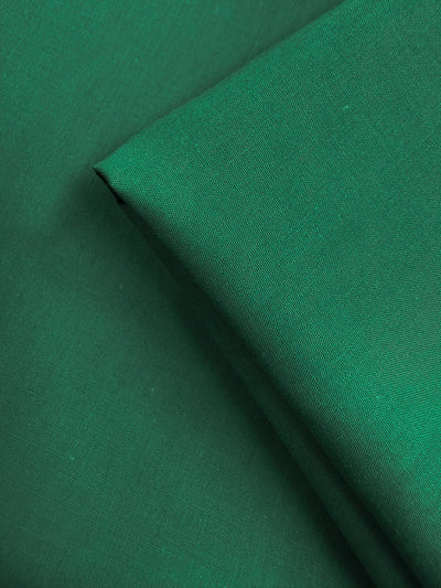 Two pieces of Super Cheap Fabrics’ Poplin - Emerald Plain (114cm) are layered, one folded diagonally. This lightweight, slightly textured fabric in emerald green is perfect for various projects.