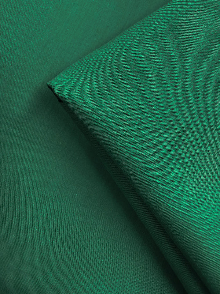 Two pieces of Super Cheap Fabrics’ Poplin - Emerald Plain (114cm) are layered, one folded diagonally. This lightweight, slightly textured fabric in emerald green is perfect for various projects.