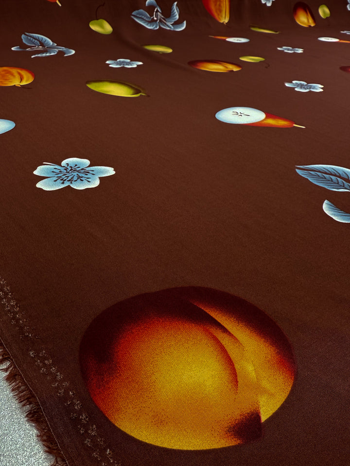 Super Cheap Fabrics' Designer Rayon - Orchard Pop - 145cm is a smooth, brown fabric with vibrant prints of oranges, pears, and apples accented by light blue flowers, all depicted in realistic colors for a lively look.