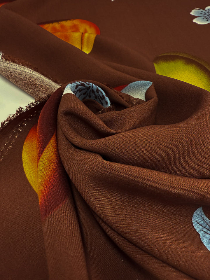 Super Cheap Fabrics' Designer Rayon - Orchard Pop (145cm) features a smooth, brown fabric with vibrant yellow, orange, and blue prints. Loosely folded, it showcases a raw, frayed edge along one side for added versatility.