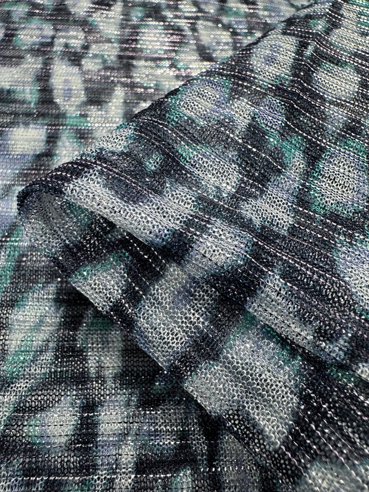 Close-up of the Metallix Pleat - Ocean Leopard fabric by Super Cheap Fabrics, showcasing a lightweight polyester blend with a folded section. It features an abstract textured pattern in shades of navy, gray, and teal, interwoven with metallic silver threads for a slightly iridescent effect. The fabric measures 140cm in width.