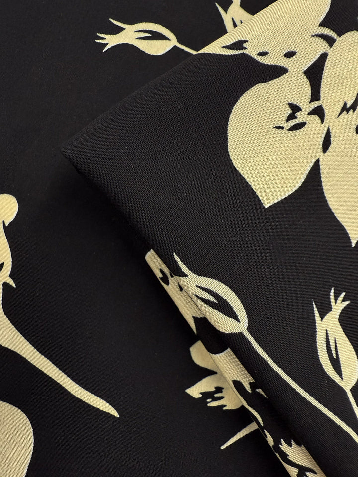 A close-up of Super Cheap Fabrics’ Designer Rayon - Moonflower Silhouette (147cm) in black with a beige floral pattern, featuring overlapping layers and stylized flower and leaf designs.