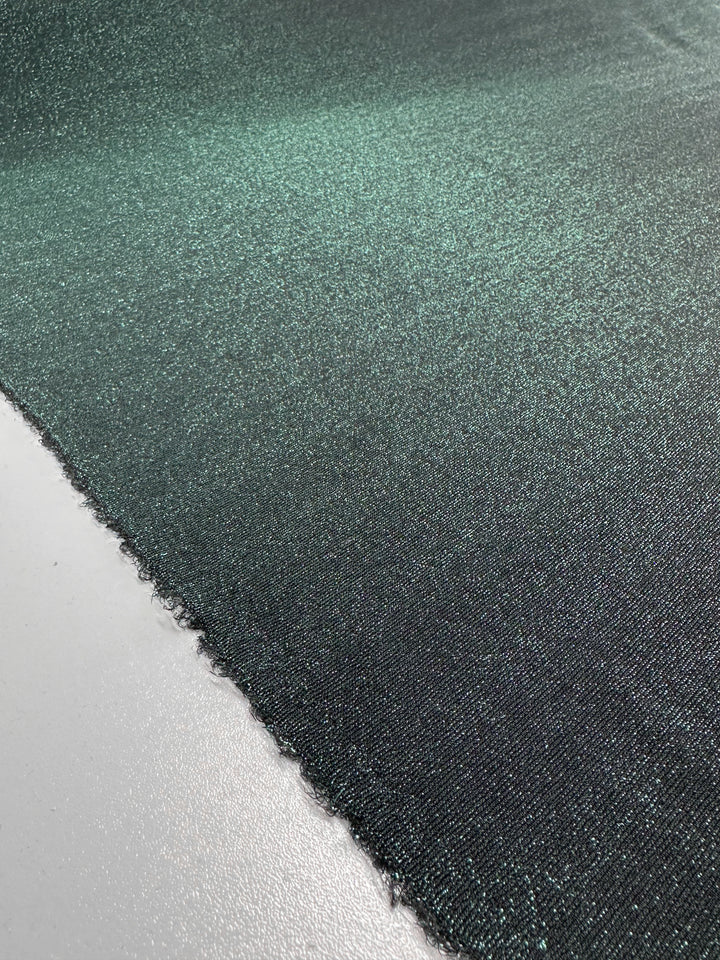 The product features a close-up of Super Cheap Fabrics' Metallic Lycra in Green, designed for jackets and measuring 170cm. The fabric is laid flat, showcasing its smooth, glossy texture with a shimmering dark green hue and a subtle metallic sheen. Raw edges can be seen on the left side, evoking the elegance typical of sophisticated dresses.