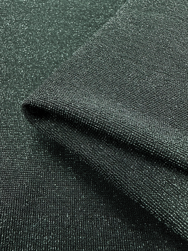 The close-up of the Folded Metallic Lycra from Super Cheap Fabrics, with its dark green hue and subtle sparkle reminiscent of a luxurious green jacket, reveals a fine weave and soft sheen. This fabric's smooth and elegant appearance makes it ideal for crafting chic dresses. With a width of 170cm, it's perfect for your next creation.