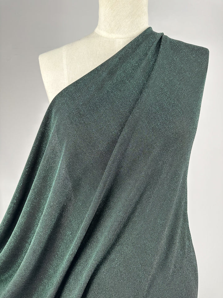 A mannequin dressed in a shimmering dark green jacket crafted from Metallic Lycra fabric by Super Cheap Fabrics, showcasing a one-shoulder style. The material has a subtle sparkle, lending texture to the piece's simple yet elegant drape. A plain, neutral backdrop enhances the jacket's allure.