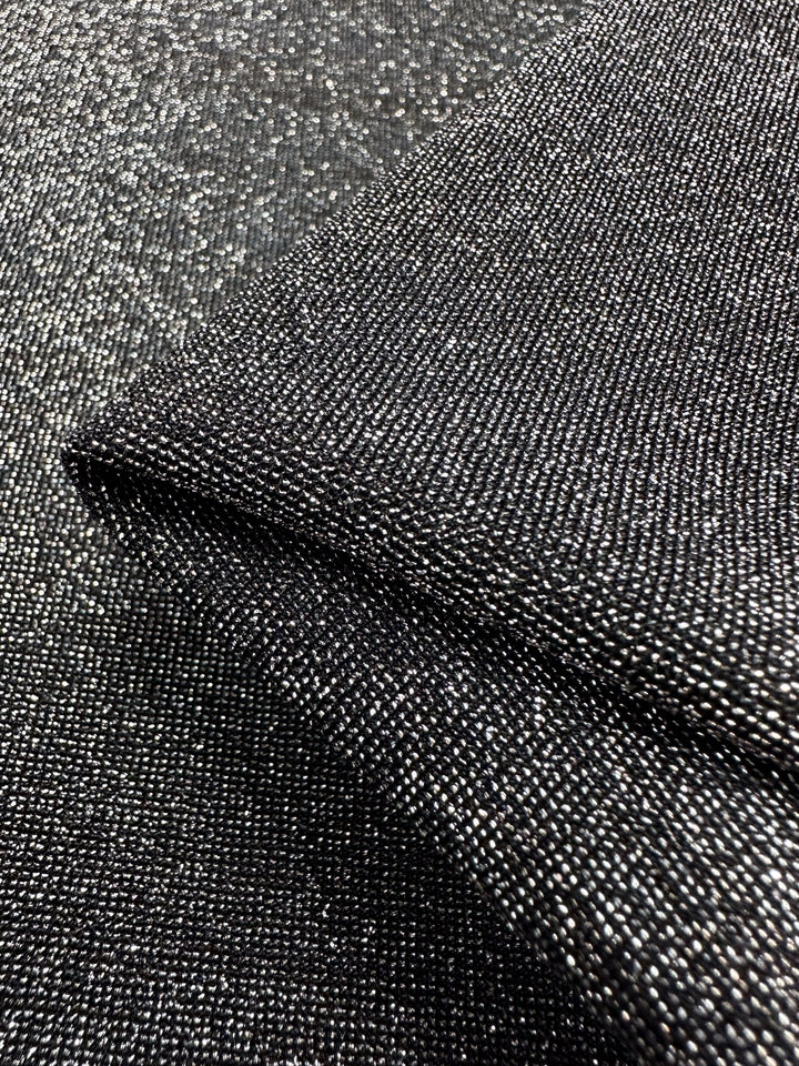 A close-up view of the Metallic Lycra - Silver - 160cm from Super Cheap Fabrics reveals its textured and shimmering metallic gray surface. The fabric is artfully folded to accentuate its intricate weave and reflective quality, making it perfect for crafting eye-catching tops or dresses.