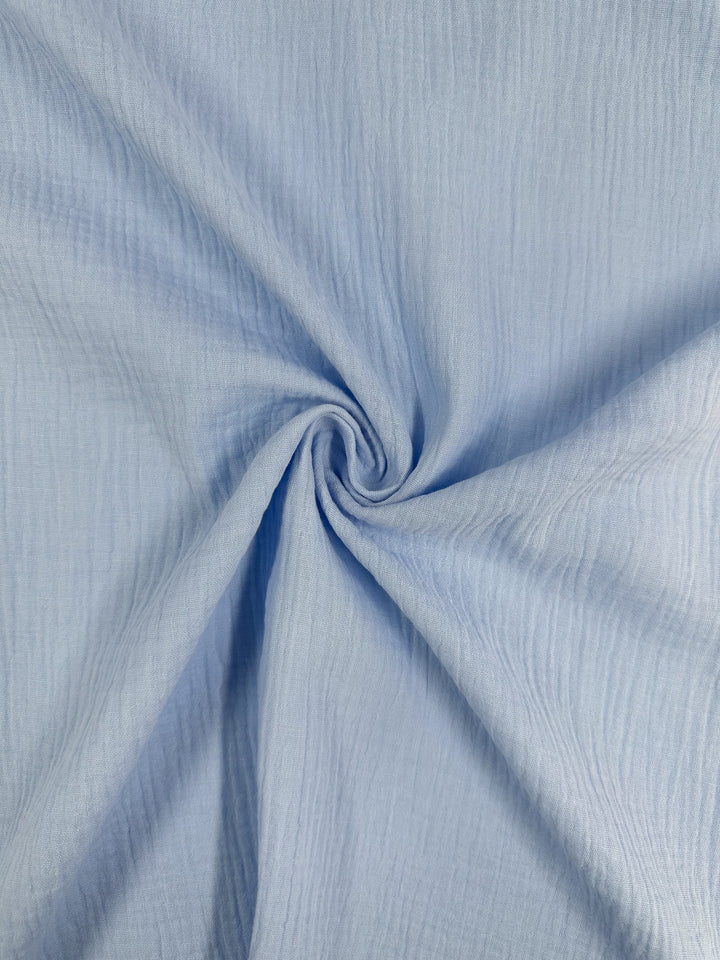 A close examination of the Serenity muslin in light blue from Super Cheap Fabrics reveals an organic fabric with a subtle textured pattern. Measuring 135cm, this material is artfully arranged in a spiral, crafting a dynamic swirl effect at the center—perfect for those who value elegance and sustainability.