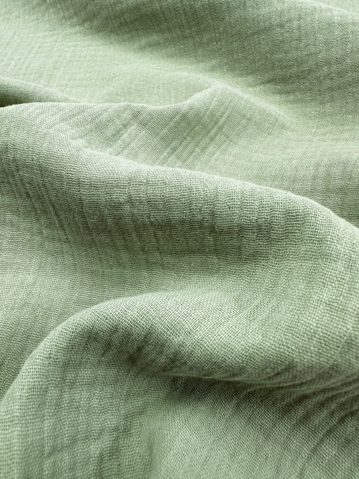 Close-up of the Muslin - Laurel Green - 135cm from Super Cheap Fabrics, featuring a soft and textured appearance with a crumpled look. This organic fabric displays a subtle woven pattern that adds depth and movement across its surface, making it perfect for various sustainable projects.