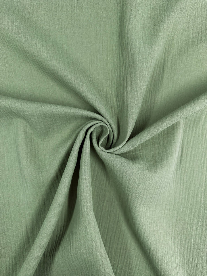 A close-up of the Super Cheap Fabrics' Muslin in Laurel Green (135cm) shows the fabric twisted at the center, creating a spiral pattern. The organic material features a textured, creased appearance that adds depth to its soft green shade, emphasizing its potential as a reusable cloth.