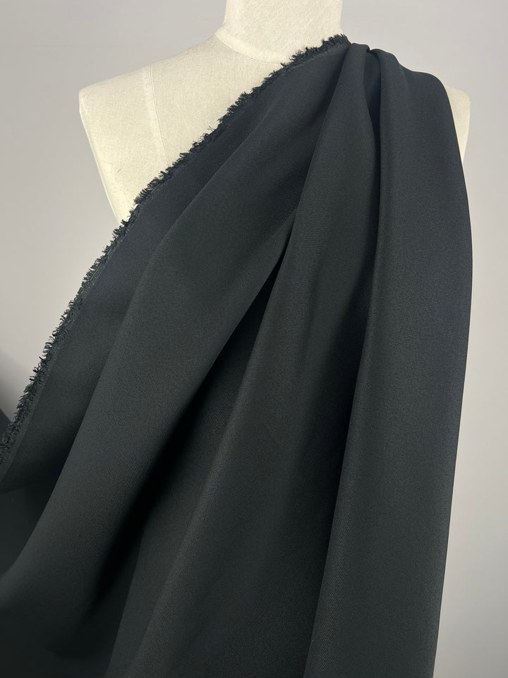 A mannequin draped in an elegant, medium weight black cotton twill fabric from Super Cheap Fabrics' Stretch Cotton Twill - Black Night - 145cm collection, with a raw, unfinished edge. The fabric flows smoothly across the mannequin, showcasing its texture and drape, set against a neutral background.