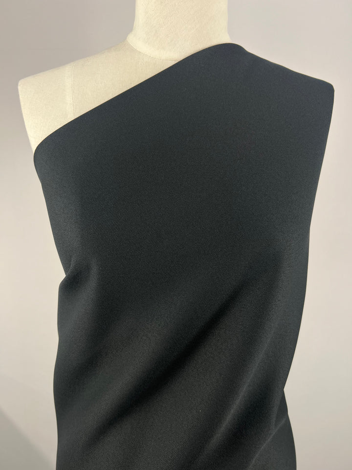 A mannequin draped in Super Cheap Fabrics' Stretch Cotton Twill - Black Night - 145cm stands against a plain background. The medium-weight fabric covers the mannequin from the left shoulder down, leaving the right shoulder bare. The simplicity of the setup highlights the texture and drape of this exquisite black twill fabric.