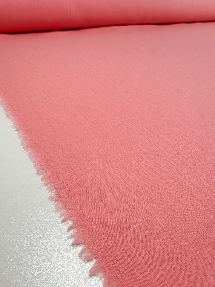 A close-up view of a roll of Super Cheap Fabrics' Muslin in Flamingo Pink, featuring a slightly textured surface and slightly frayed edges. This organic fabric roll is placed on a flat, light-colored surface.