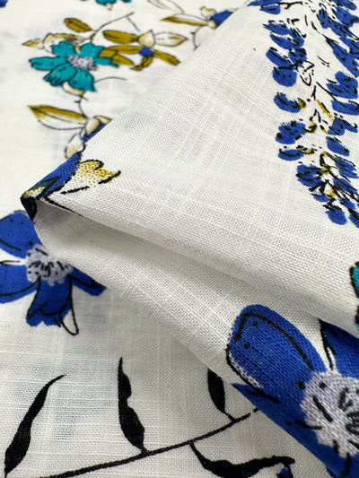 A close-up of "Linen Blend - Royal Vine - 145cm" by Super Cheap Fabrics showcases a breathable white linen fabric adorned with a vibrant floral pattern. This lightweight blend features intricate blue, green, and yellow flowers accompanied by black leaves, making it an ideal choice for colorful summer outfits.