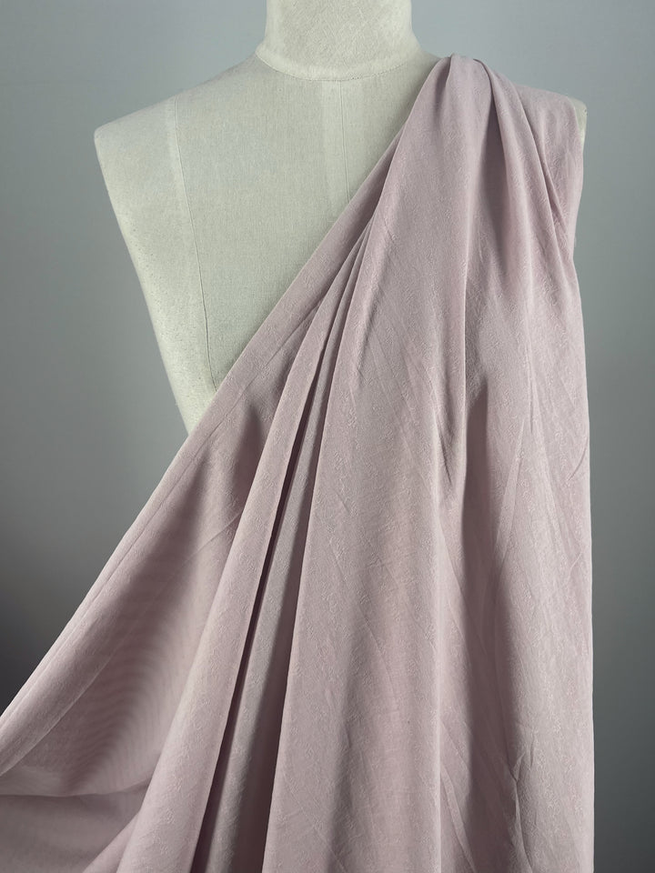 A graceful drape of Textured Cotton in Burnish Lilac from Super Cheap Fabrics elegantly cascades over a white mannequin, highlighting the material's smooth texture and gentle folds. The extra lightweight fabric stands out beautifully against a plain gray background.