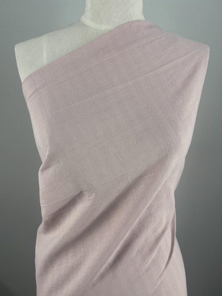 A mannequin is draped with Super Cheap Fabrics' Textured Cotton in Burnish Lilac, its 100% cotton composition revealed through a single exposed shoulder. This extra lightweight fabric's delicate hue and subtle sheen are enhanced against the plain gray background, vividly displaying its texture.