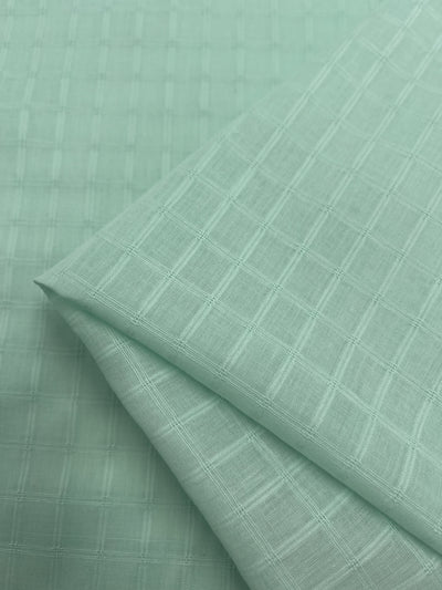 A close-up view of Super Cheap Fabrics' Textured Cotton - Honeydew - 145cm showcases its extra lightweight, light green fabric with a grid pattern. The material is neatly folded, revealing the intersecting lines that form the subtle, repeating checkered design.