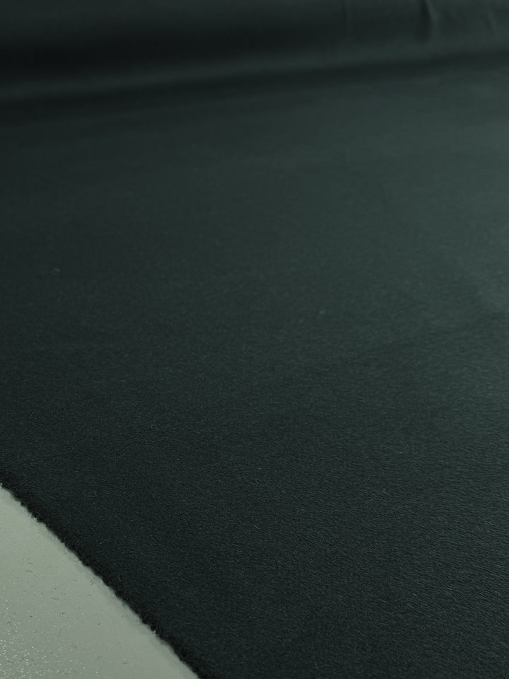 A close-up of Super Cheap Fabrics' Wool Cashmere - Sea Moss - 146cm shows the large, flat, dark green luxury fabric laid out smoothly, with soft lighting revealing its rich texture.