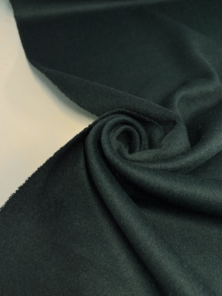 A close-up of Super Cheap Fabrics' Wool Cashmere - Sea Moss (146cm) shows its dark green, soft, and slightly fuzzy texture, loosely twisted in a spiral against a light background.