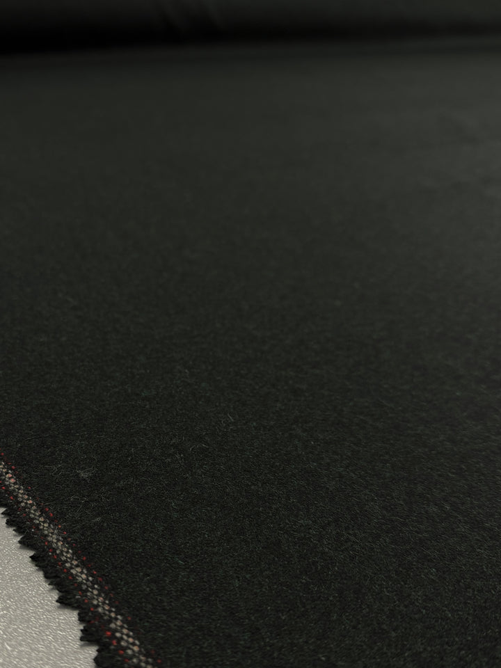 A close-up of Super Cheap Fabrics' Melton Wool - Rosin Marle (150cm) shows the dark, smooth heavyweight wool with stitched edge, lying flat. The stitching lines the left side while the fabric extends into the background.