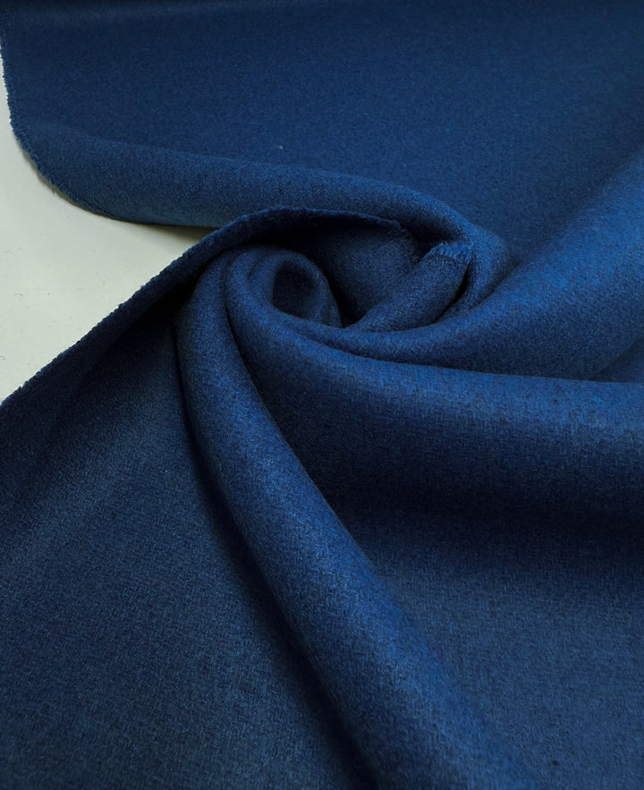 A close-up of Super Cheap Fabrics’ Melton Wool - Blue Quartz - 150cm, a heavyweight outerwear fabric in deep blue, loosely twisted on a flat surface to highlight its soft texture, smooth finish, and impressive thickness.