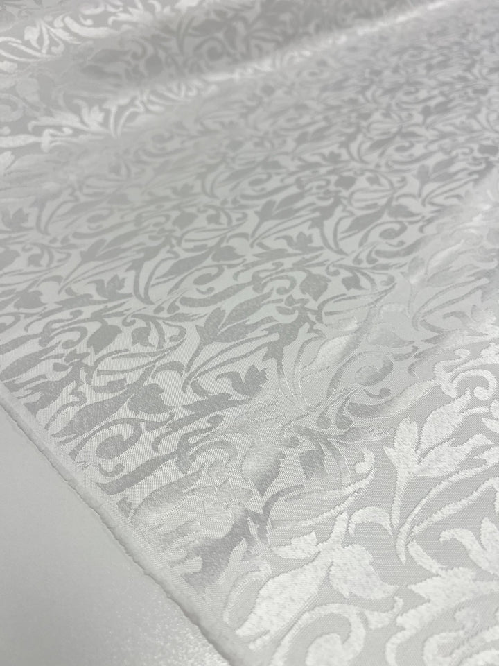 A close-up of the Designer Damask - White - 155cm from Super Cheap Fabrics showcases an intricate floral pattern, with swirling leaves and vines that contribute to its elegant, textured appearance on the smooth fabric surface.