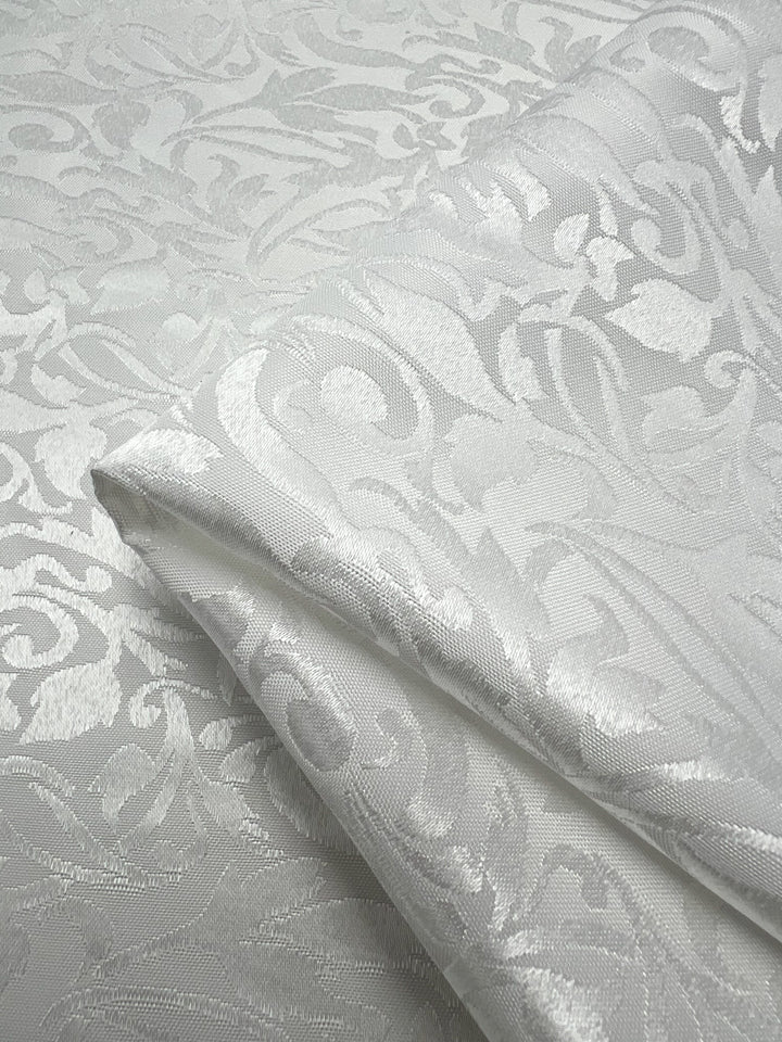 A detailed close-up of the Designer Damask fabric by Super Cheap Fabrics showcases its luxurious, intricately woven floral and leaf motifs. The white 155cm fabric's subtle sheen accentuates its sophisticated design, elegantly displayed against a matching patterned backdrop.