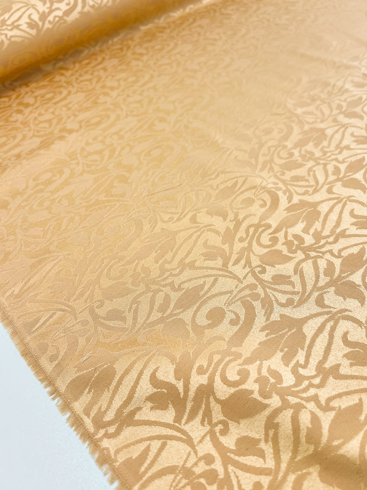 A close-up of the Designer Damask - Oak Buff - 155cm from Super Cheap Fabrics reveals an intricate, swirling floral pattern in a luxurious gold hue. The silky texture and subtle shine beautifully showcase the detailed design across the material.