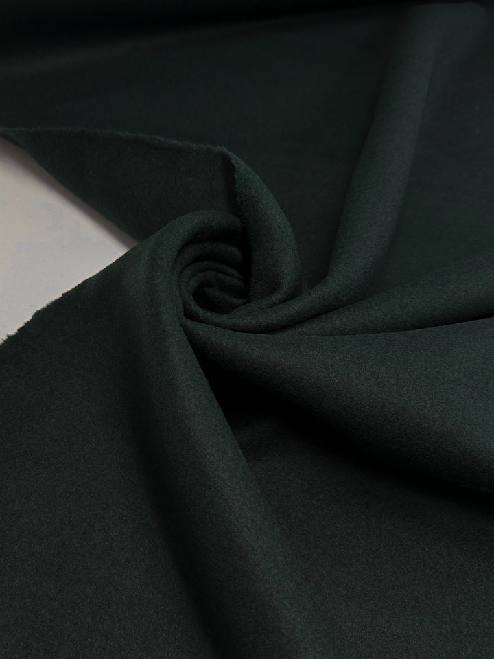 A close-up of Super Cheap Fabrics' Melton Wool - New Scarab (154cm) reveals its dark green, heavyweight, and smooth texture, elegantly twisted to showcase drape and flexibility—ideal for luxurious outerwear.