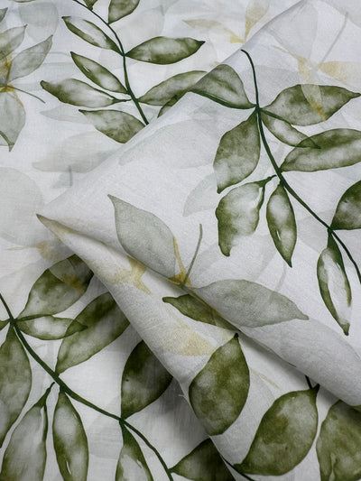 A close-up of the Designer Cotton - Gully - 150cm by Super Cheap Fabrics, showcasing its lightweight cotton fabric adorned with a leafy green pattern. The varying shades of leaves create a natural and fresh aesthetic, perfect for luxury sewing projects. Portions of the fabric are folded, adding depth and texture to the image.