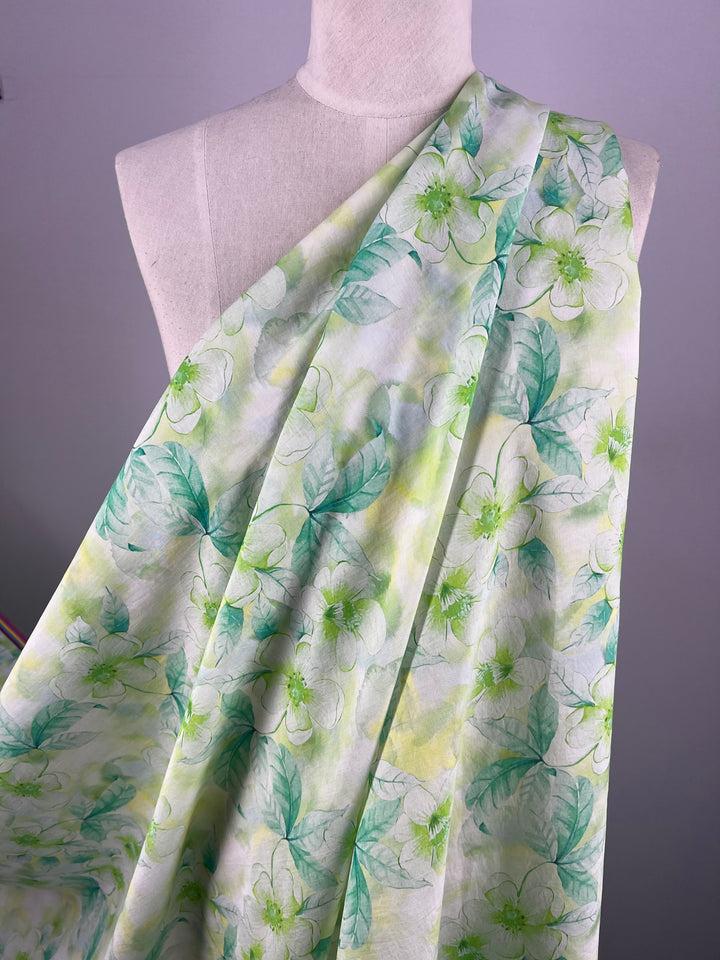 A white mannequin draped with Super Cheap Fabrics' Designer Cotton - Periwinkle - 150cm featuring a floral pattern. The fabric is white with green and yellow leaves, and large light green flowers interspersed throughout. Perfect for luxury sewing projects, the scene is set against a plain, light-colored wall.