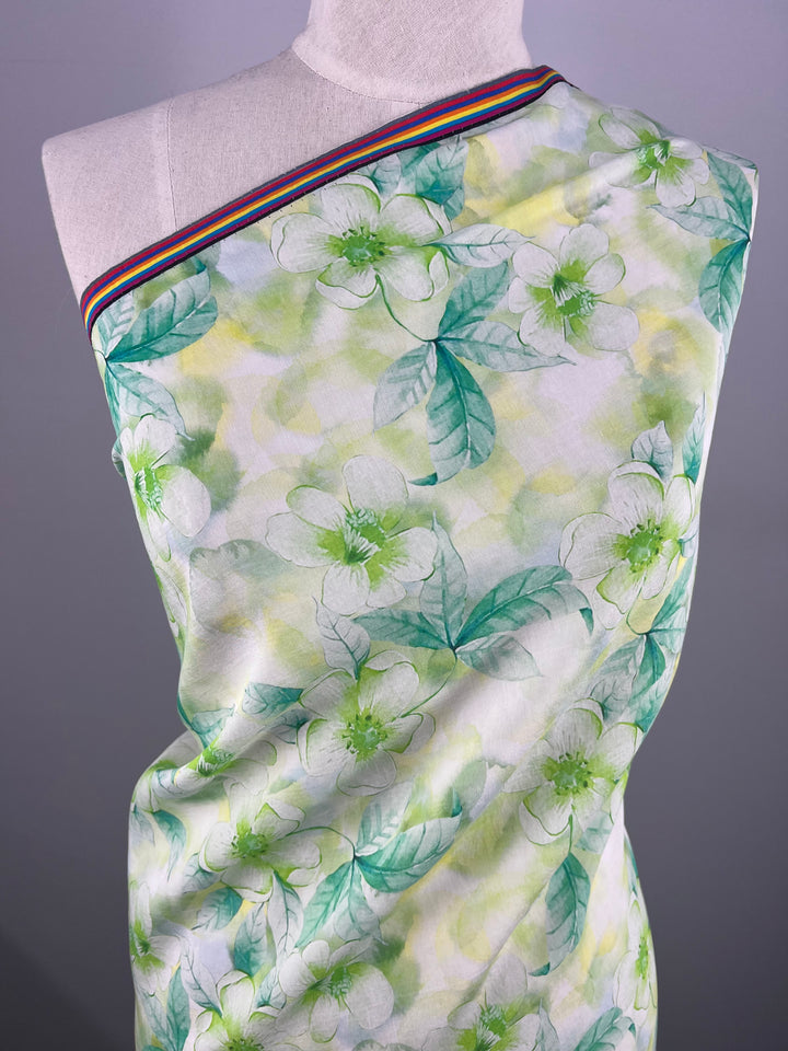 A dress form is adorned with Super Cheap Fabrics' Designer Cotton in Periwinkle, featuring a light green and white floral pattern with green leaves. The breathable fabric is draped over one shoulder and has a multicolored striped ribbon along the neckline. The background is a plain gray, perfect for luxury sewing projects.