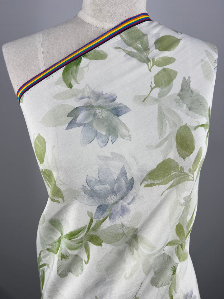 A white fabric from Super Cheap Fabrics, named Designer Cotton - Lotus - 150cm, elegantly draped over a dress form. It features a green and lavender floral pattern with large flowers and leaves. The top of this luxurious fabric is accented with a narrow rainbow stripe running along the upper edge, making it perfect for premium sewing projects.