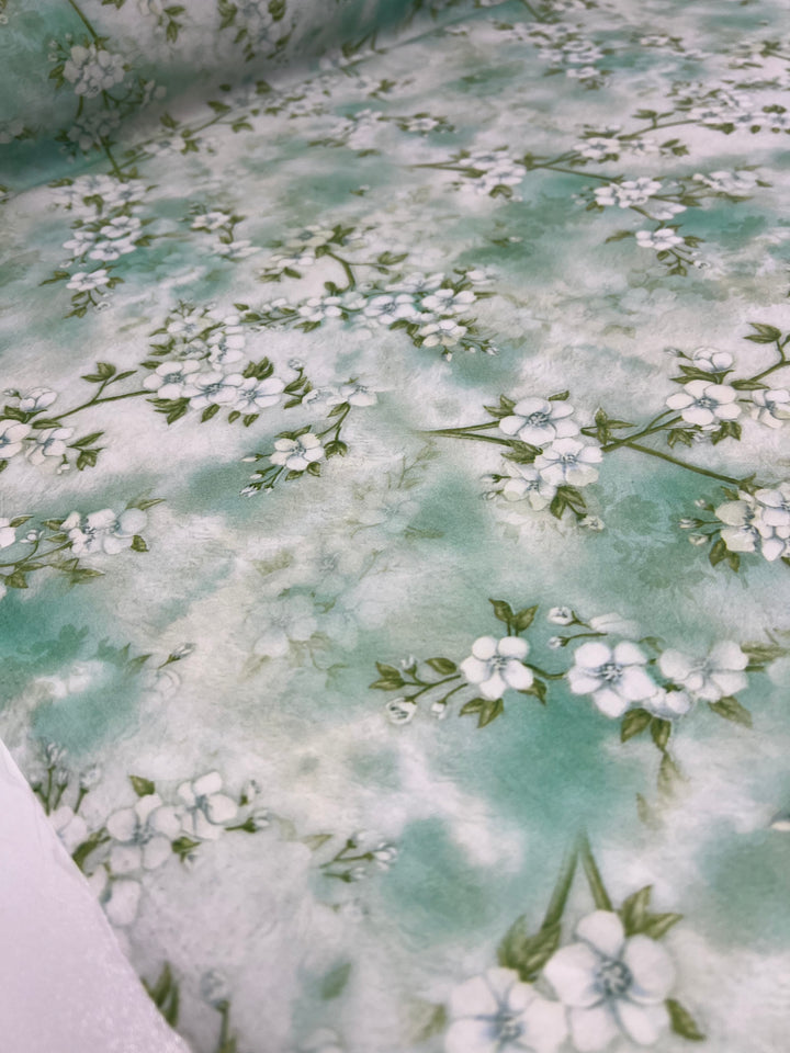 A close-up of a fabric roll showcases the "Designer Cotton - Blossom" by Super Cheap Fabrics, featuring a light green and white floral pattern. The fabric boasts a watercolor effect background with green stems and white flowers scattered across it, creating a soft, delicate design perfect for luxury sewing projects using this premium designer cotton. The roll measures 150cm in width.