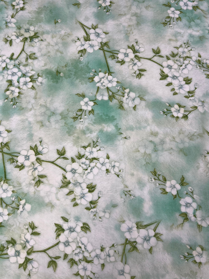 Introducing the "Designer Cotton - Blossom - 150cm" by Super Cheap Fabrics, a luxurious soft designer cotton fabric adorned with delicate white blossoms and green leaves scattered across a gentle gradient background of light blue and white. This exquisite floral pattern evokes a springtime feel, making it perfect for luxury sewing projects.