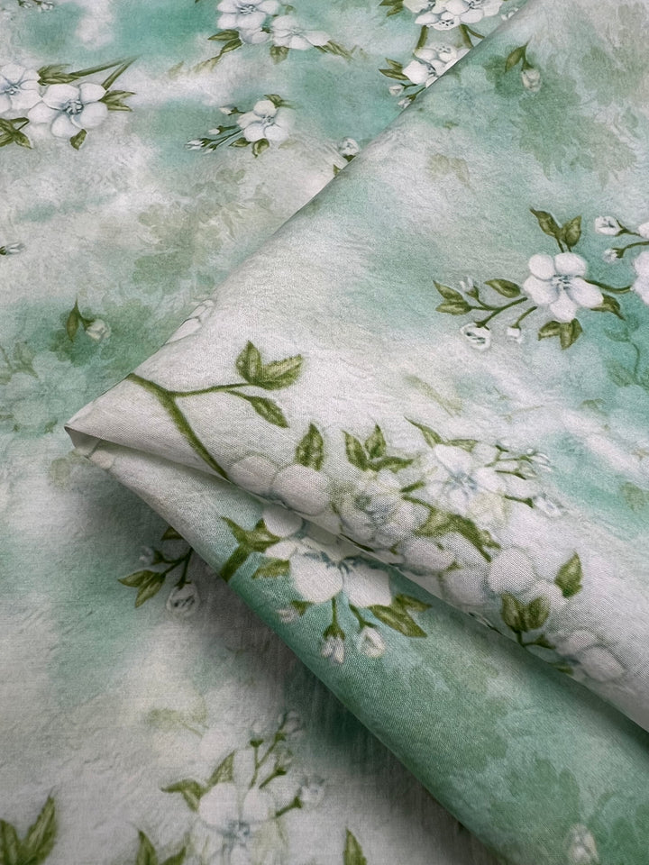 The Designer Cotton - Blossom by Super Cheap Fabrics showcases a sophisticated soft green fabric with a delicate white floral pattern, highlighted by light green leaves. The fabric is neatly folded, emphasizing the seamless floral design against a gradient background that transitions from light to darker green hues, making it ideal for luxury sewing projects. This high-quality cotton fabric measures 150cm in width and is perfect for creating elegant and stylish pieces.
