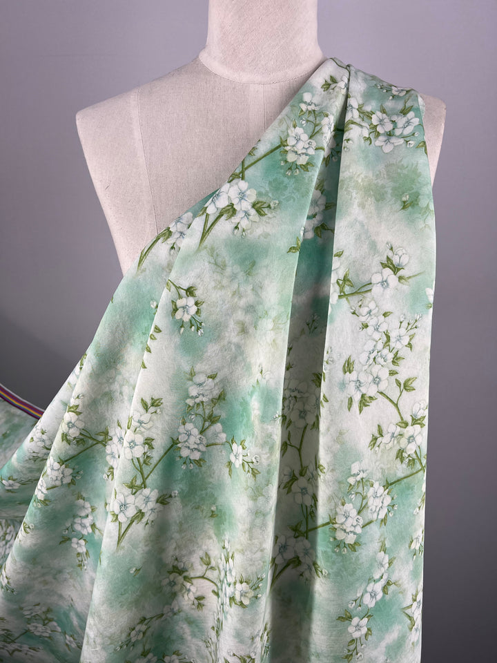 A mannequin showcases a piece of fabric draped over one shoulder. The fabric, ideal for luxury sewing projects, boasts a light green background adorned with small, delicate white flowers and green stems. Crafted from designer cotton under the name "Designer Cotton - Blossom - 150cm" by Super Cheap Fabrics, it contrasts beautifully against a neutral gray backdrop.