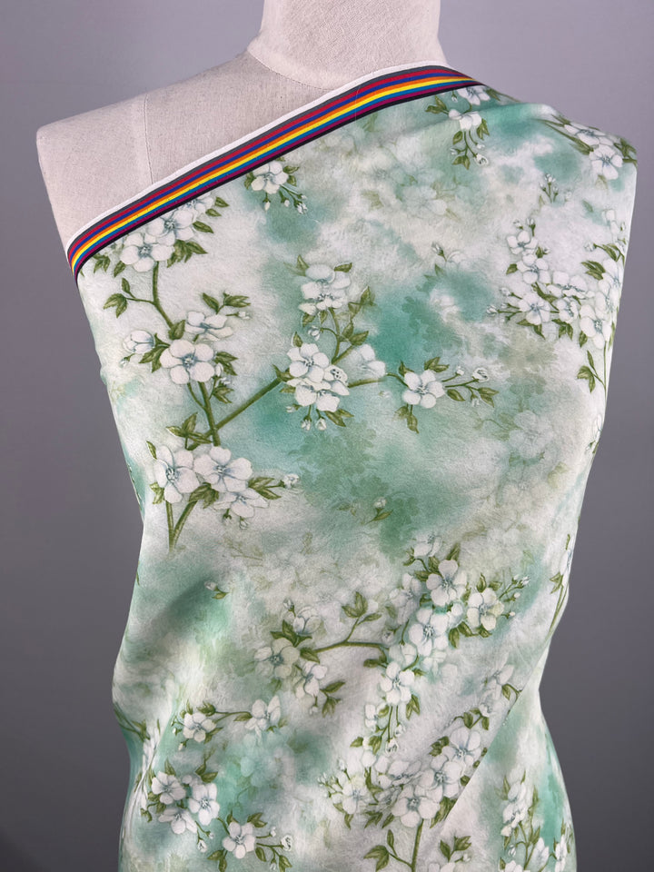 A mannequin is draped in Super Cheap Fabrics' Designer Cotton - Blossom, a light green fabric adorned with white floral patterns. The breathable material is styled off-shoulder, showcasing a rainbow-striped border along the top edge. The neutral background highlights the intricate design, making it perfect for luxury sewing projects.